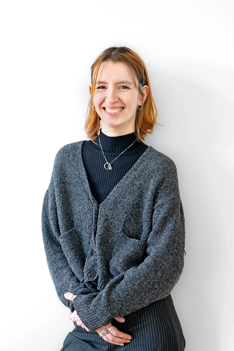 Paula steht angelehnt und lächlnd an einer weißen Wand.